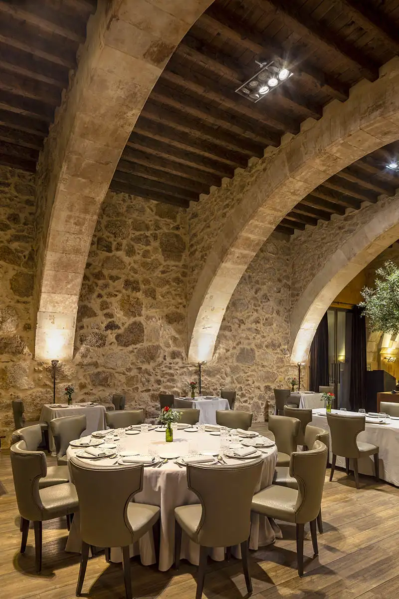 Comedor con mesa puesta y sillas junto a columnas de piedra en sal&oacute;n con techo de vigas.