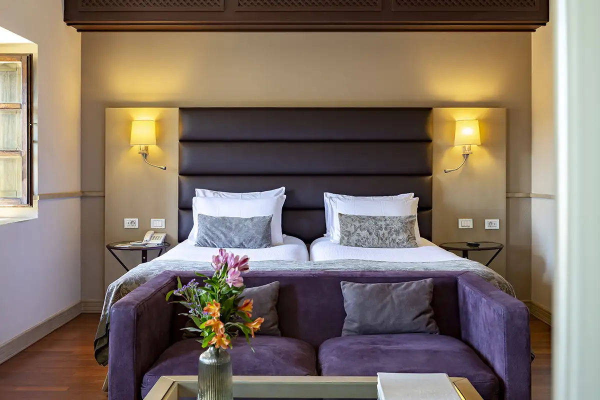 Habitaci&oacute;n de hotel con cama doble, cojines grises y flores en jarr&oacute;n