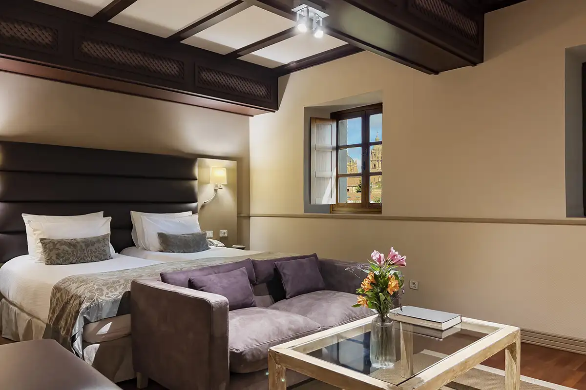 Suite hotelera moderna con sof&aacute;, cama y ventana a vista de campanarios