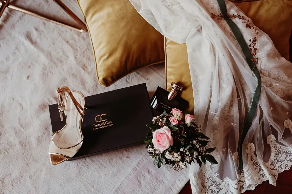 Zapatos de tac&oacute;n, caja y perfume junto a un ramo de rosas sobre tela.