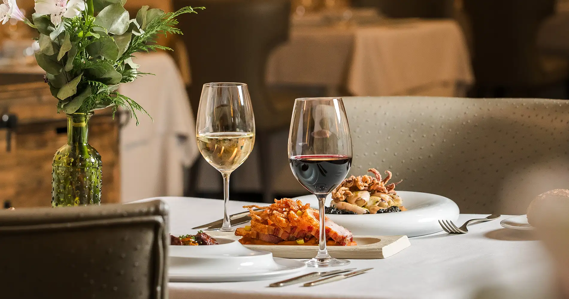 Mesa con dos copas de vino y platos servidos, con botella y flores al fondo