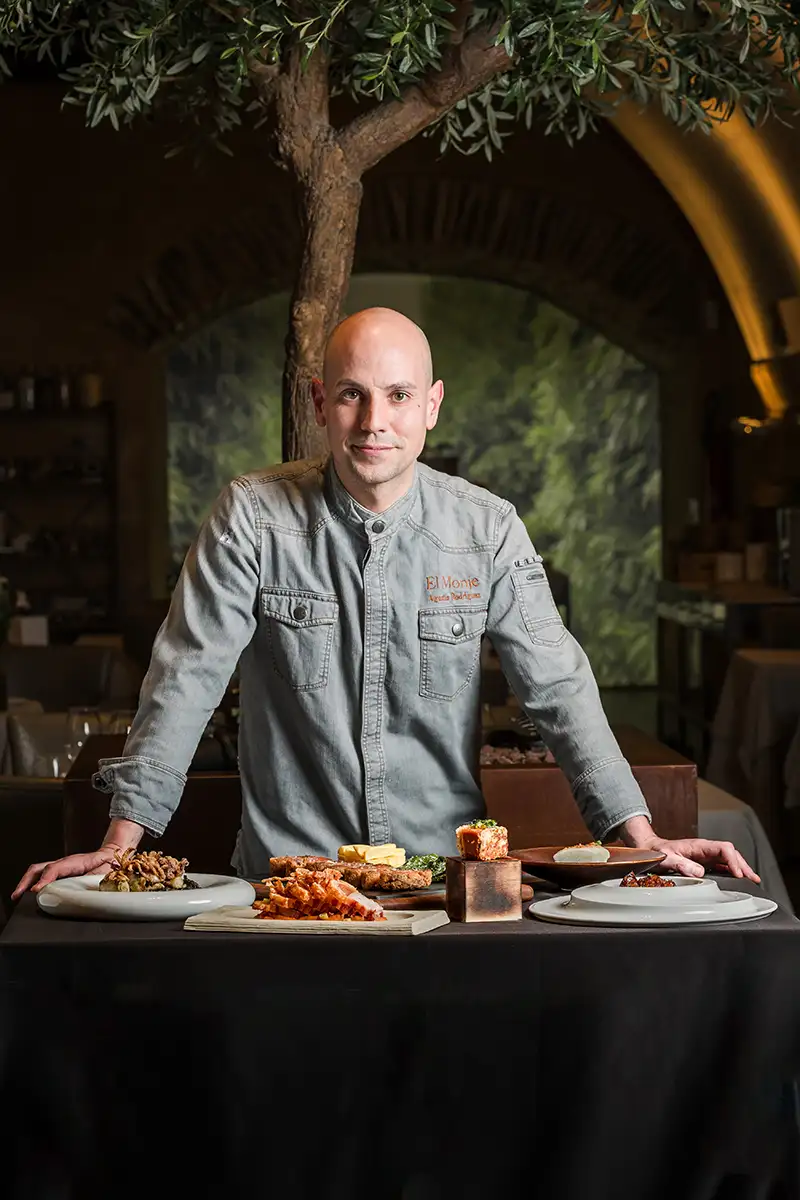 Chef con chaqueta gris sentado frente a mesa con platos y comida.