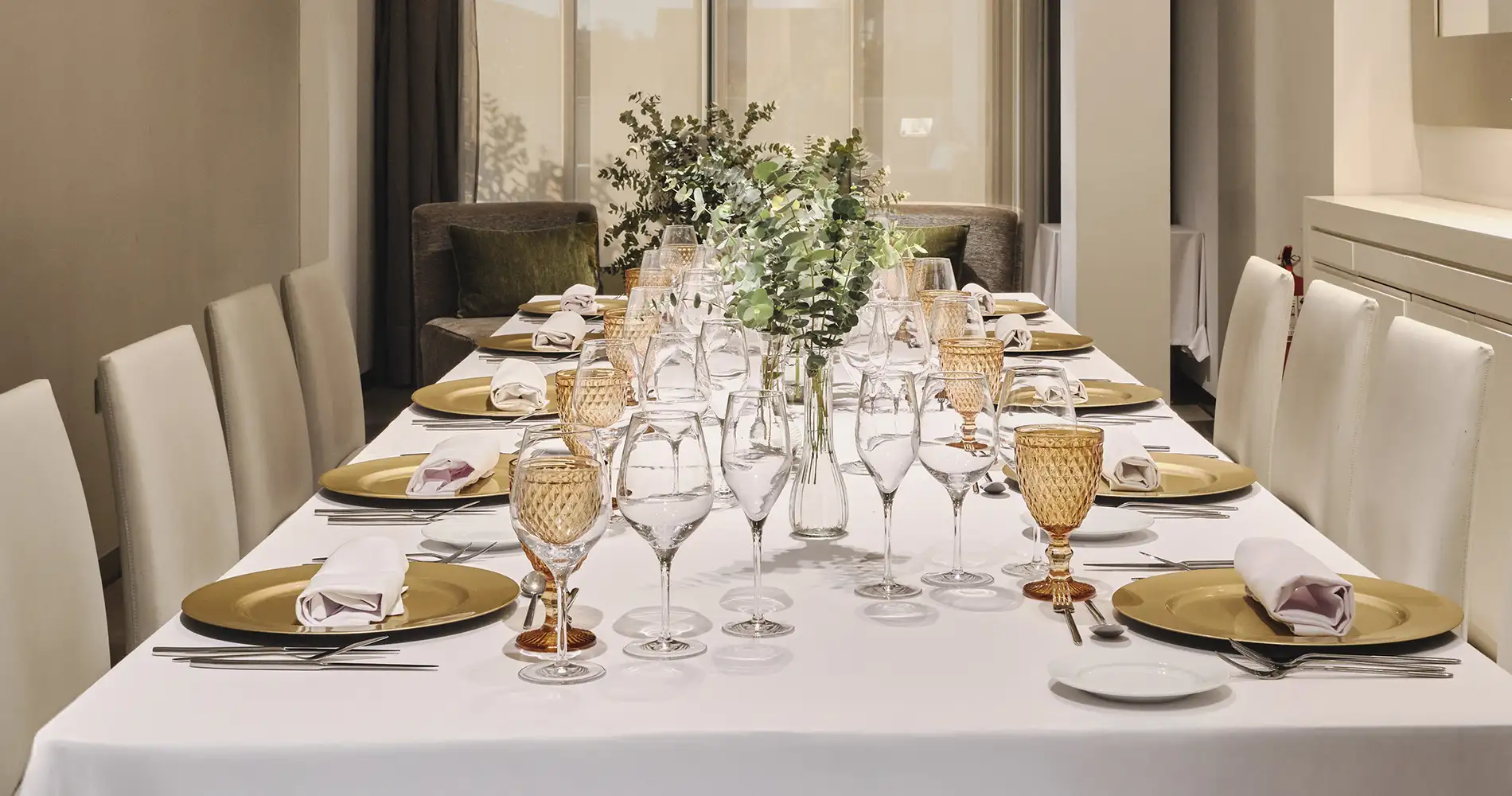 Mesa de comedor elegante con copas, platos dorados y arreglo de eucalipto