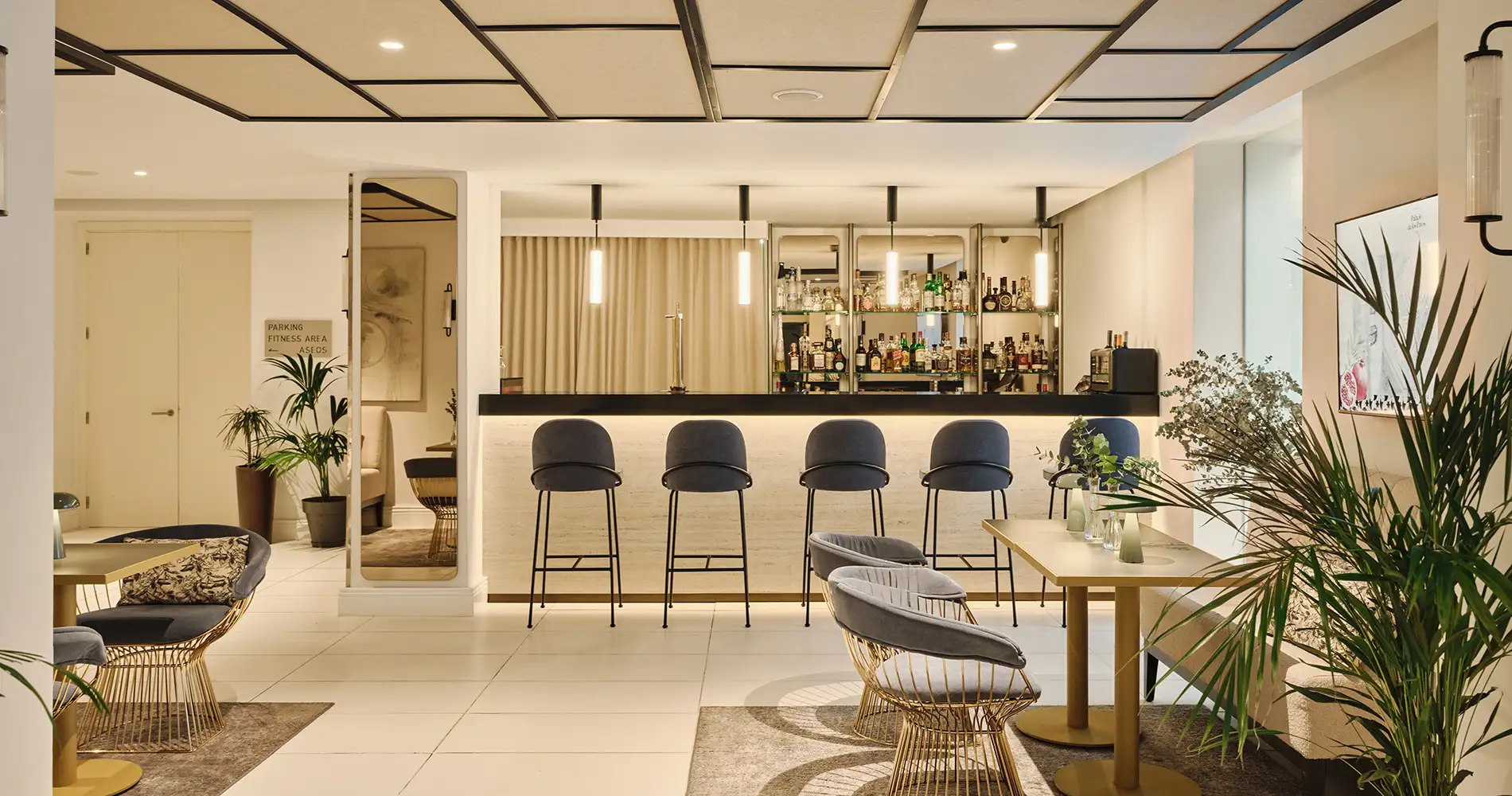 Bar con botellas, sillas y mesas, iluminaci&oacute;n interior c&aacute;lida, plantas.