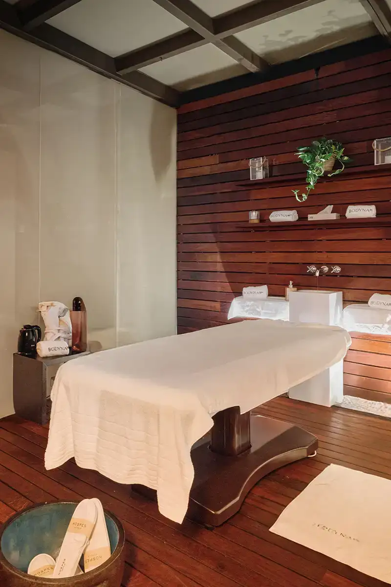 Sala de masaje con camilla blanca y pared de madera con productos y plantas