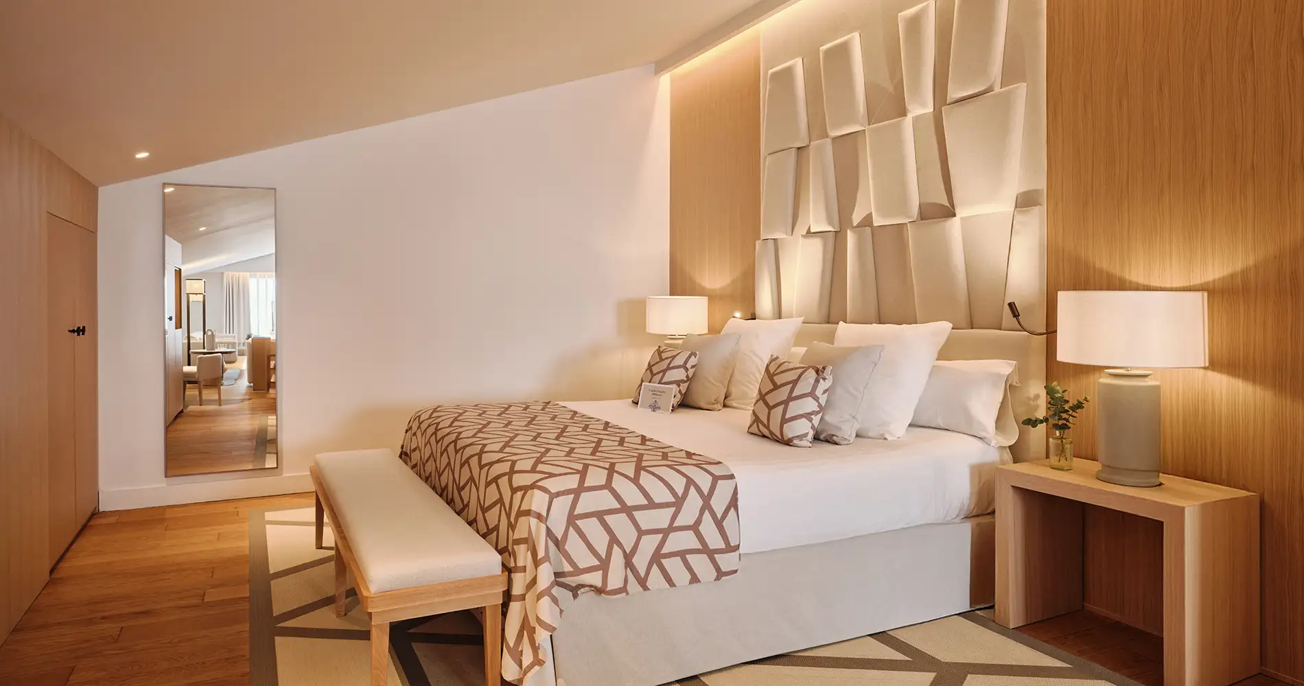 Dormitorio moderno con cama, almohadas y l&aacute;mparas junto a espejo y pared decorativa
