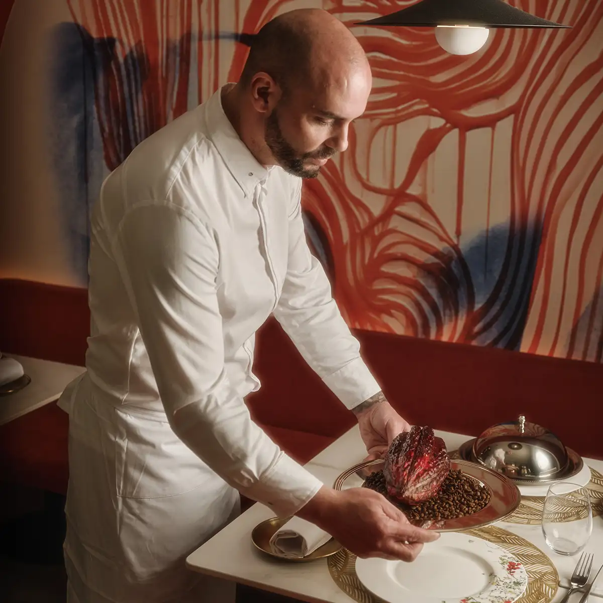 Chef con gorra blanca sirve carne sobre plato en mesa con mural rojo