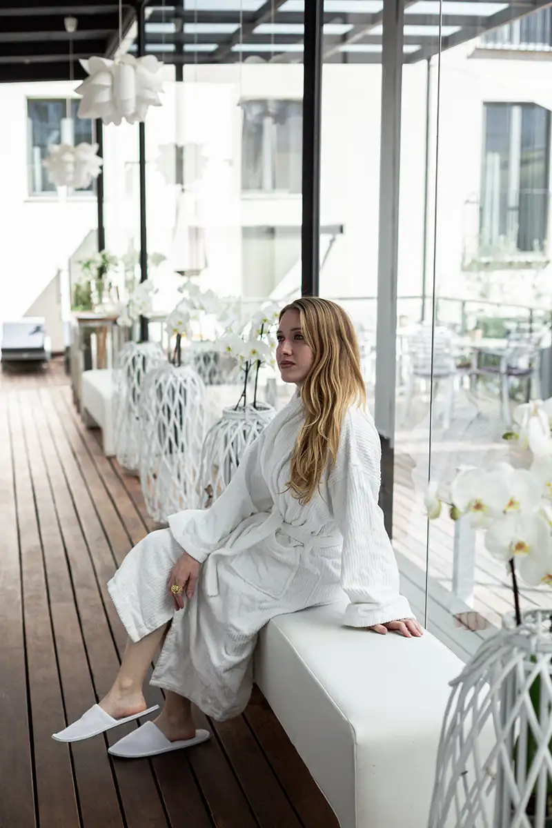 Mujer con bata blanca sentada en terraza con ventanales y flores blancas