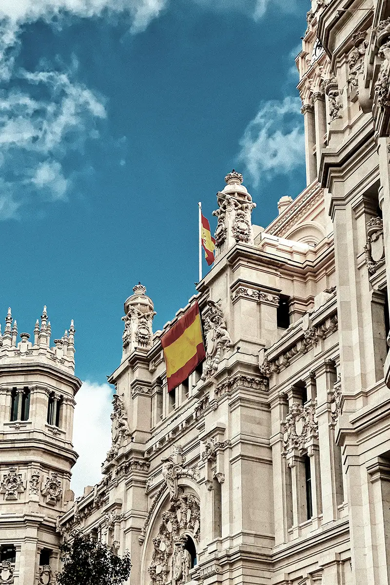 Fachada de edificios con banderas de Espa&ntilde;a y cielo azul con nubes.