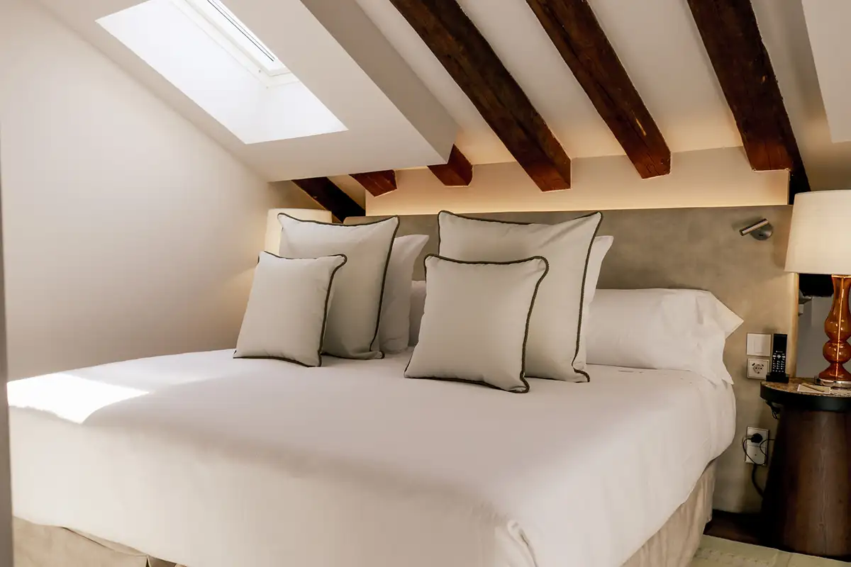 Dormitorio con cama blanca, cojines grises y vigas de madera en el techo.