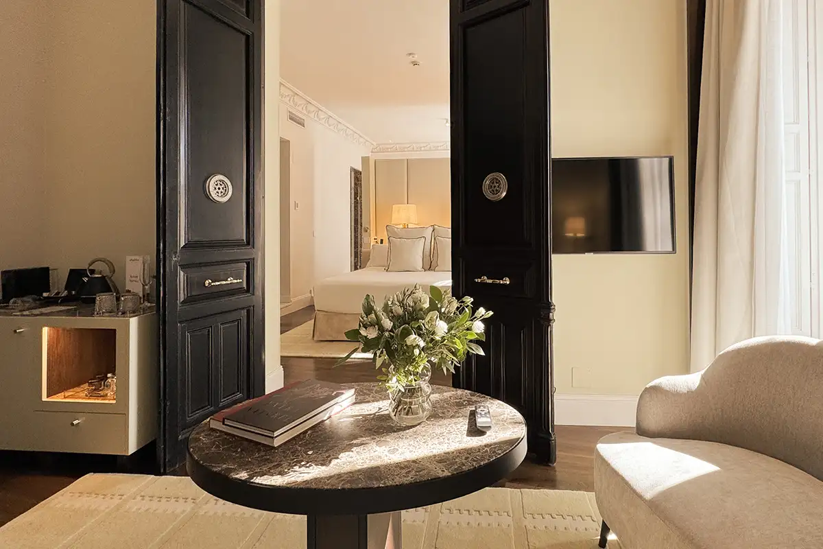 Mesa redonda con flores, sill&oacute;n beige y vista a dormitorio desde una puerta abierta.