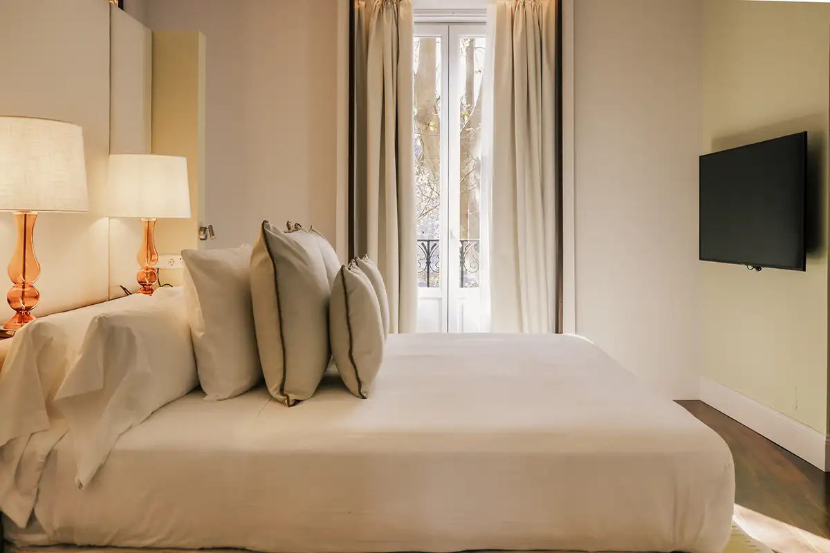 Dormitorio moderno con cama blanca, l&aacute;mparas doradas y cortinas beige.