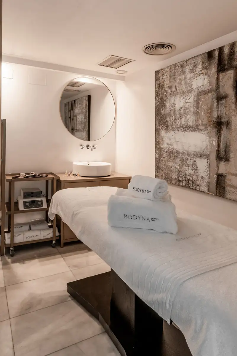 Sala de spa con cama blanca, espejo circular y lavabo, pared con textura.