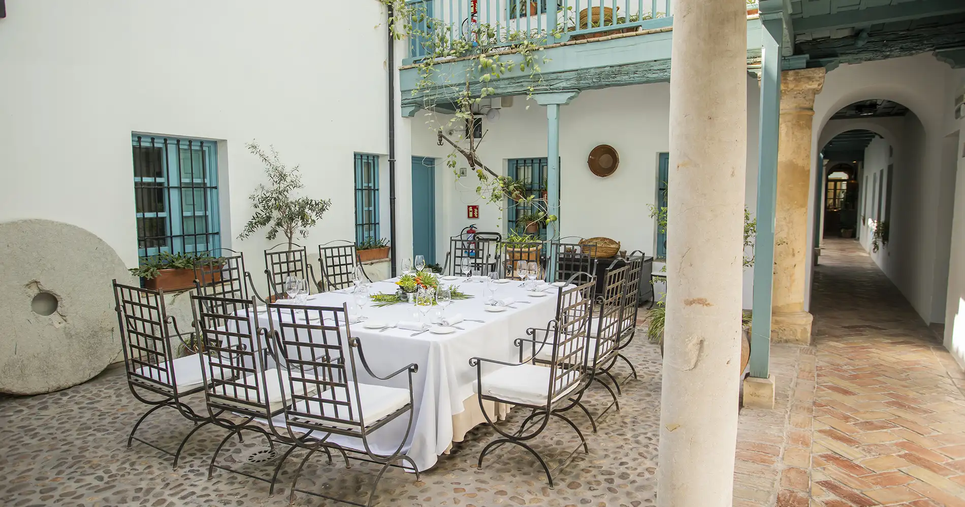 Mesa con sillas met&aacute;licas y decoraci&oacute;n floral en patio andaluz, con plantas colgantes
