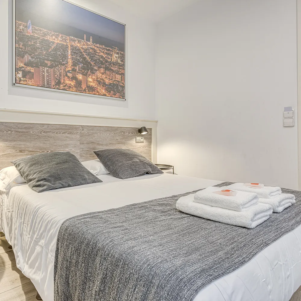 Cama doble con colchas grises, dos almohadas, toallas dobladas y cuadro en la pared.