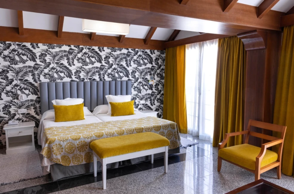 Habitaci&oacute;n doble con cama, cortinas amarillas y papel pintado tropical.