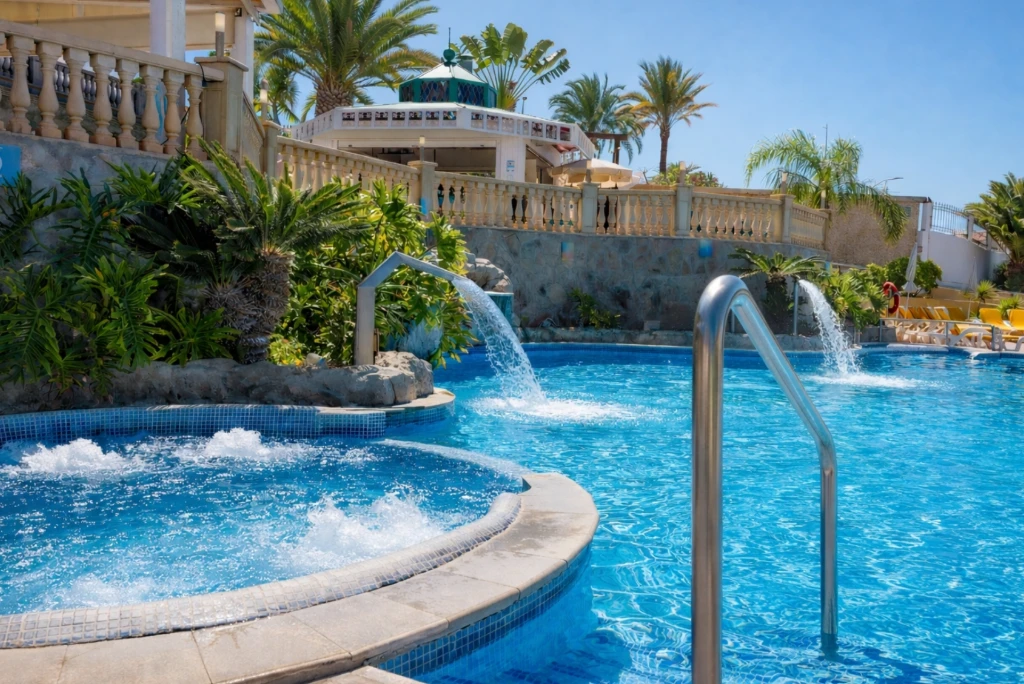 Piscina con cascadas y barandilla de acero, rodeada de palmeras y tumbonas.