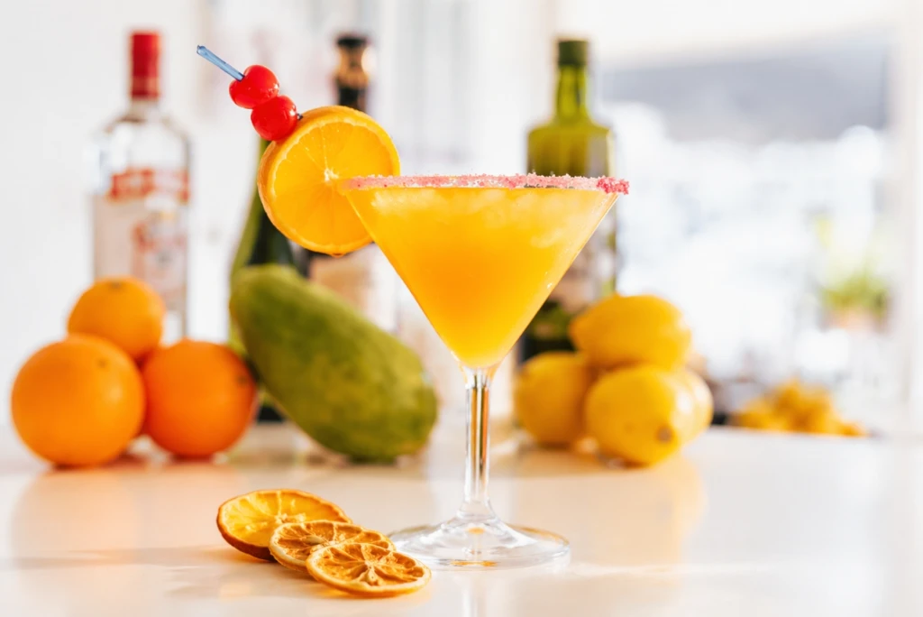 C&oacute;ctel amarillo con rodaja de naranja, borde azucarado y aceitunas decorativas.