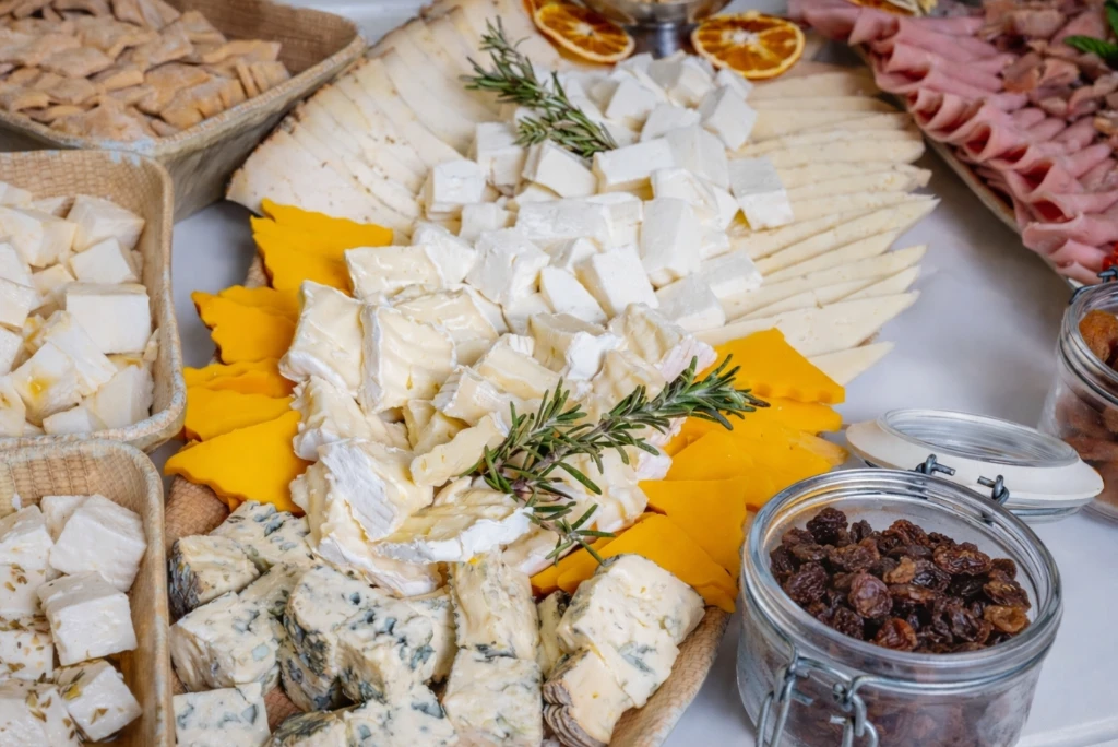 Mesa con quesos variados, hierbas y pasas en frasco, con pan y jam&oacute;n al fondo