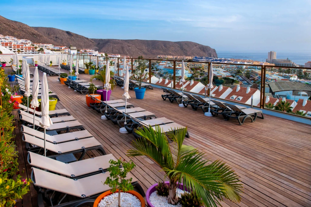 Terraza amplia con tumbonas y plantas, con vista a ciudad y mar, bajo un cielo despejado.