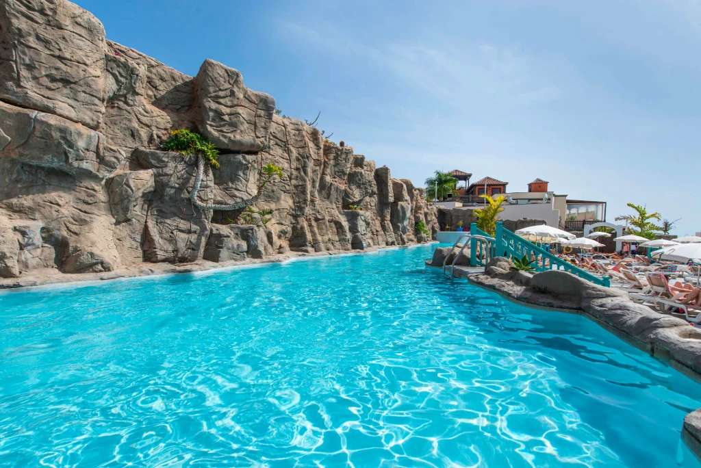 Piscina exterior con rocas artificiales y tumbonas bajo sombrillas en un d&iacute;a soleado.