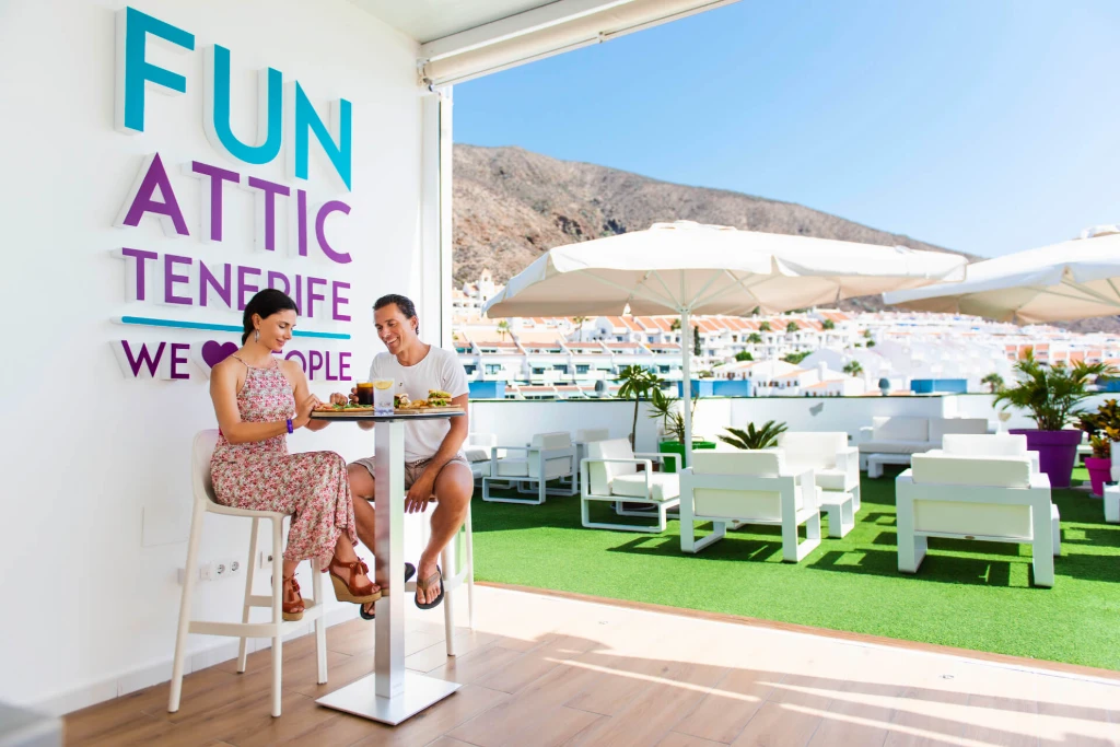 Pareja desayunando en una terraza con vista a un paisaje monta&ntilde;oso y letrero de "Fun Attic Tenerife".