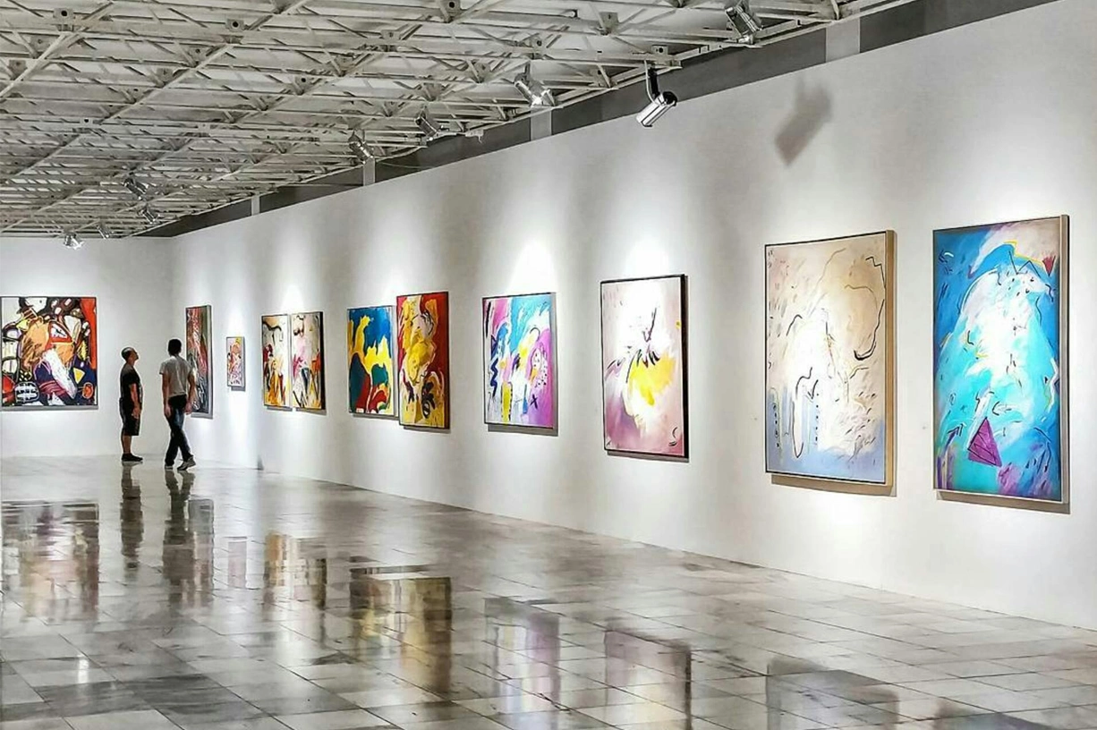 Modern art gallery interior with colorful paintings displayed on white walls and visitors walking through the space.