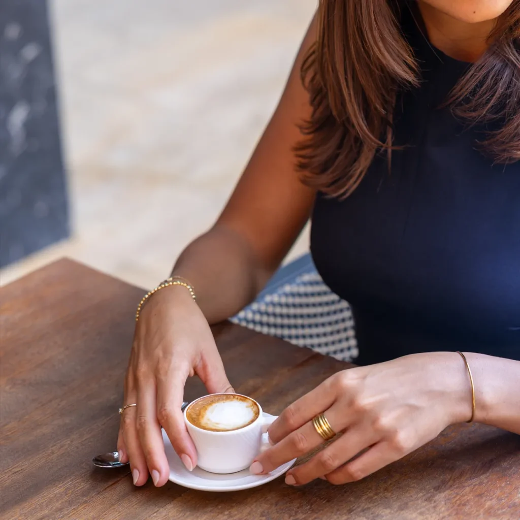 A close-up of a woman's hands holding a small cup of coffee on a wooden table.