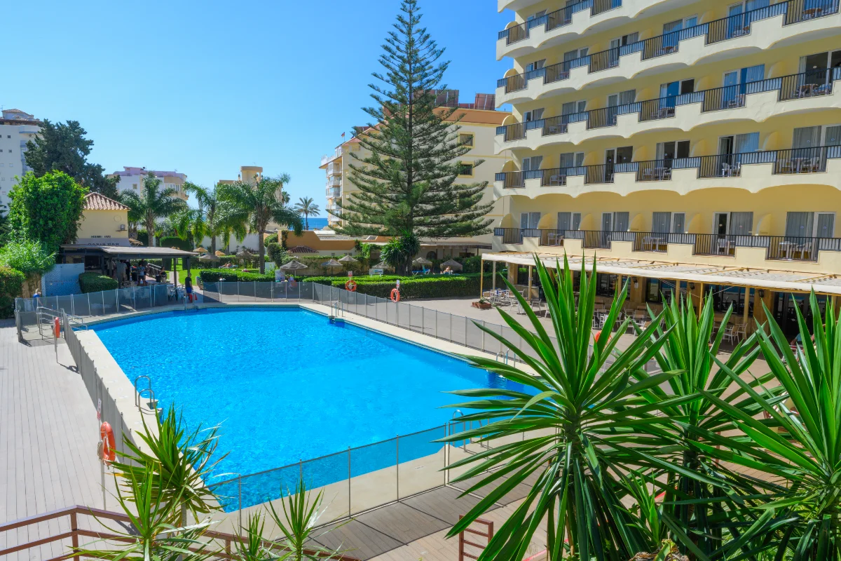 Piscina azul y edificio hotelero con balcones, rodeado de plantas y palmeras