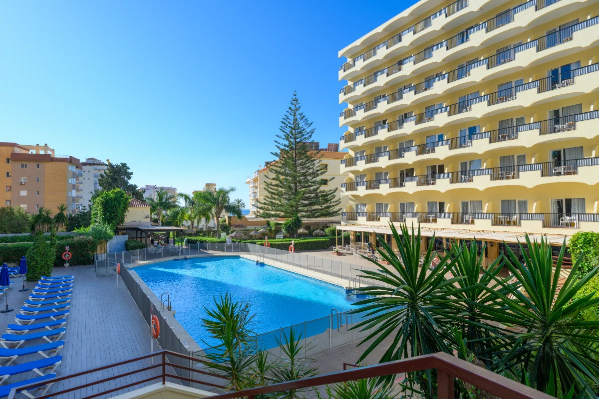 Piscina azul junto a edificio amarillo con balcones y plantas, bajo cielo despejado