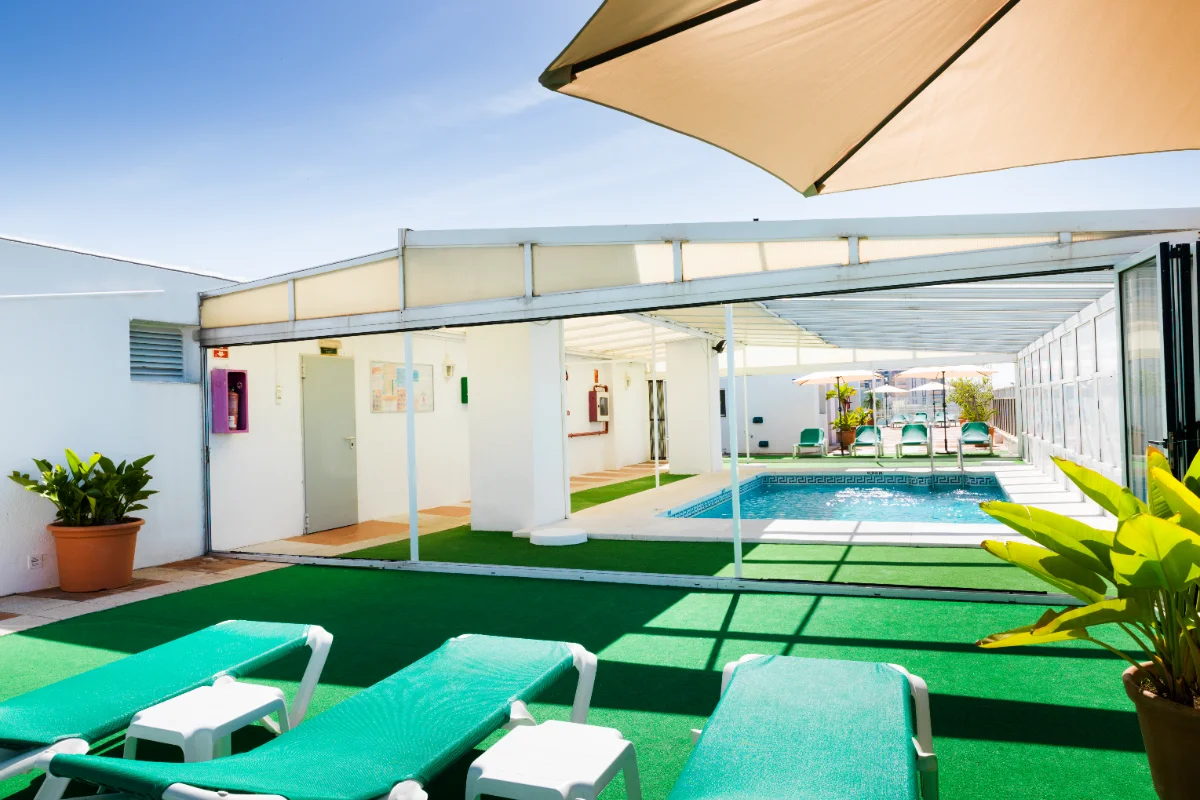 Patio con c&eacute;sped artificial, tumbonas y piscina bajo sombrilla, al aire libre