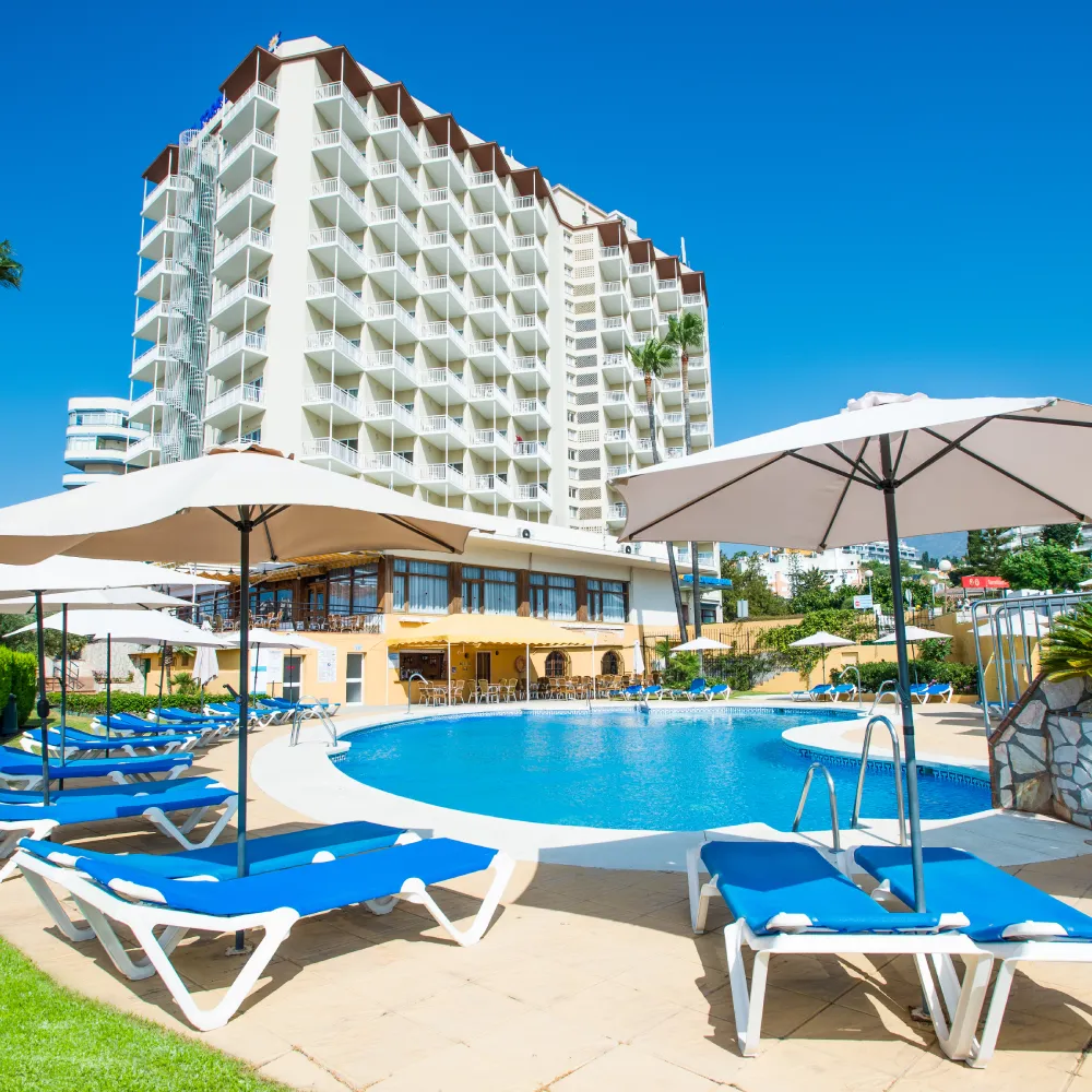 Piscina con tumbonas y sombrillas frente a un hotel de apartamentos alto