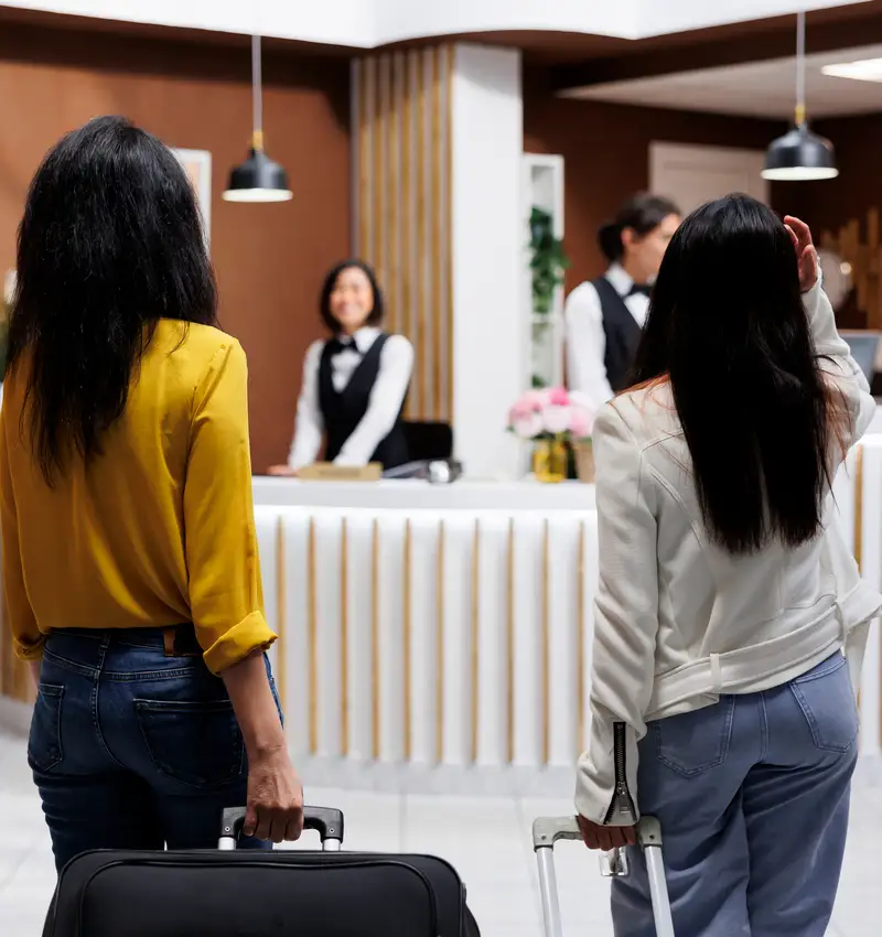 Dos mujeres en recepci&oacute;n de hotel con maletas, atentas al personal.