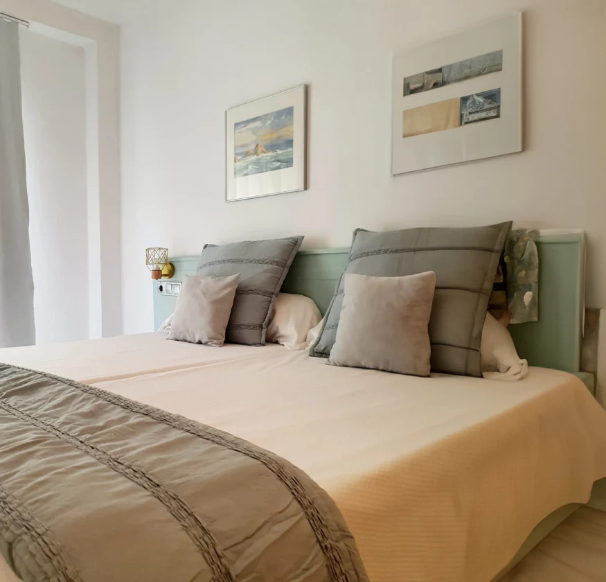 Cama doble con cojines grises y cuadros en la pared de una habitaci&oacute;n luminosa.