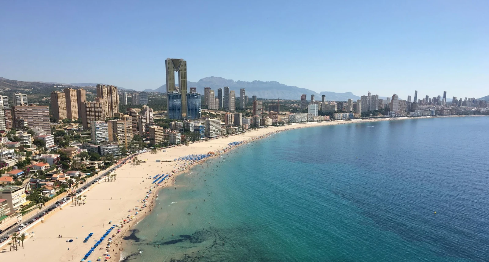 Playa con edificios altos y monta&ntilde;as al fondo bajo un cielo despejado.