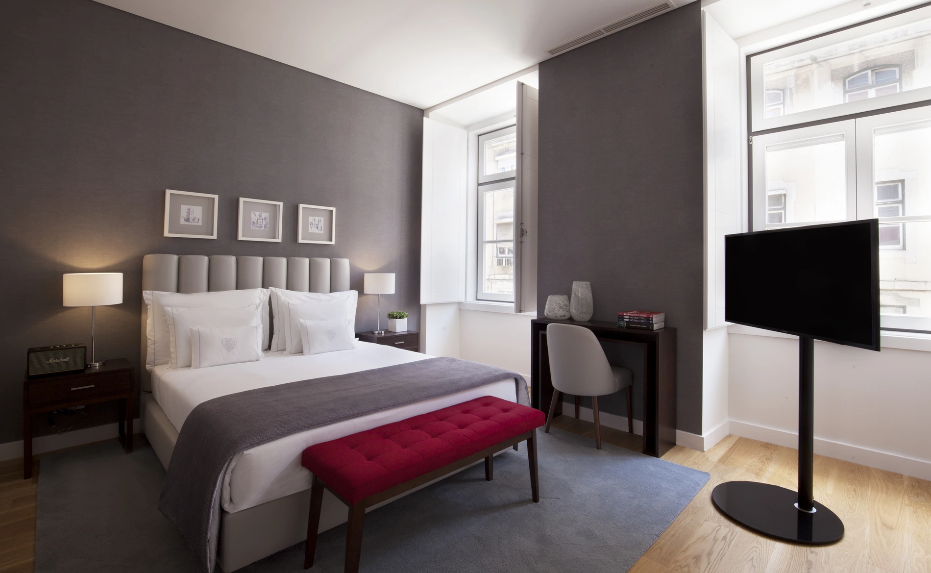 Quarto moderno com cama, mesa, TV e janela grande.