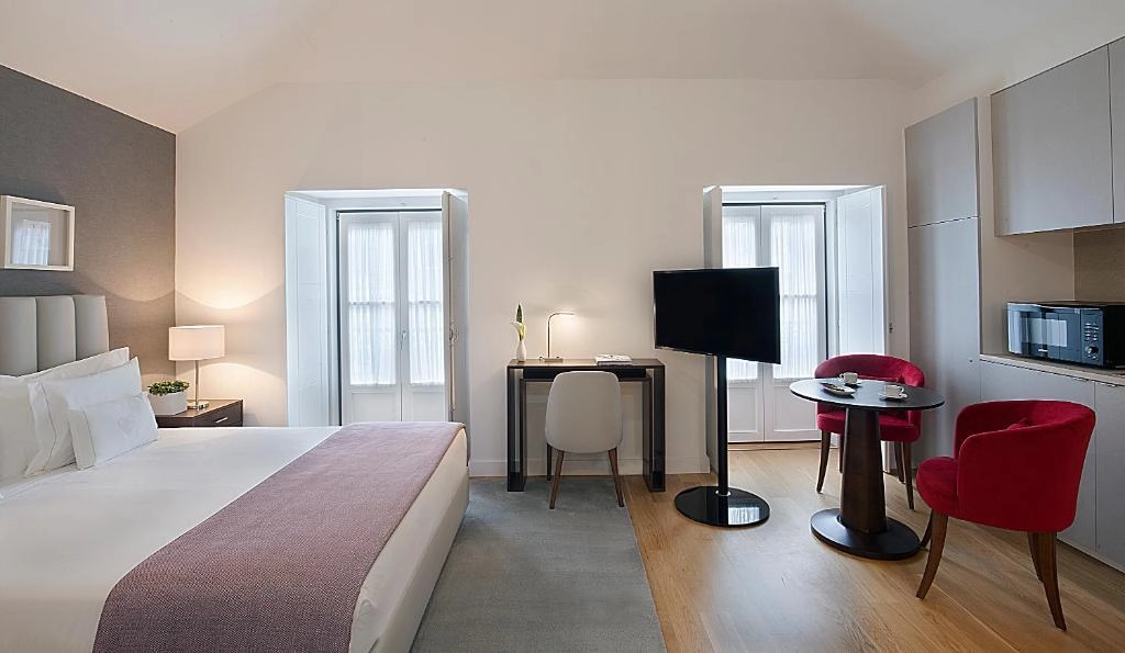 Quarto moderno com cama, mesa, TV, cadeiras vermelhas e micro-ondas.