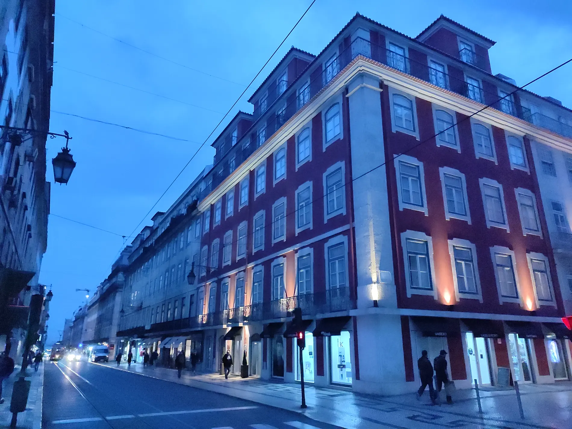 Pr&eacute;dio iluminado &agrave; noite em rua movimentada, sob c&eacute;u azul.