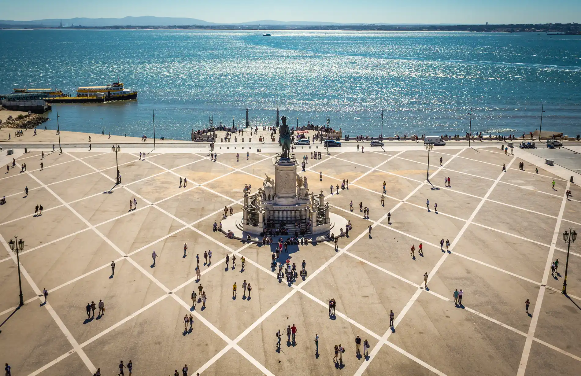Pra&ccedil;a ampla com monumento central e pessoas, ao fundo um rio e um barco amarelo.