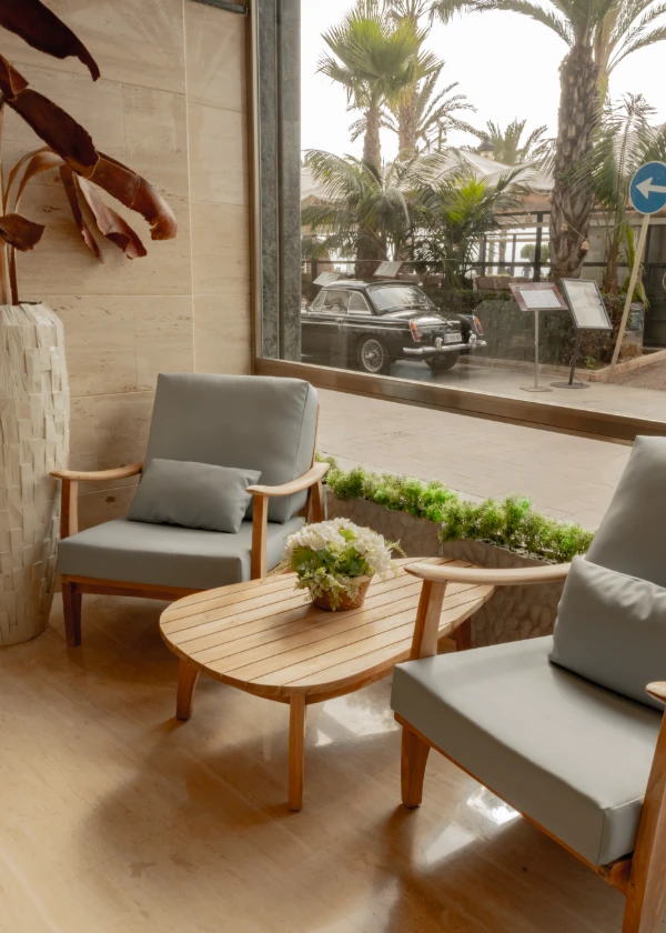 Sala con sillones y mesa de madera, ventana a la calle con palmeras y coche negro antiguo afuera.