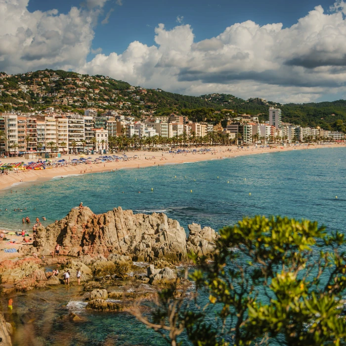Playa concurrida con edificios y colinas verdes al fondo, cielo parcialmente nublado.