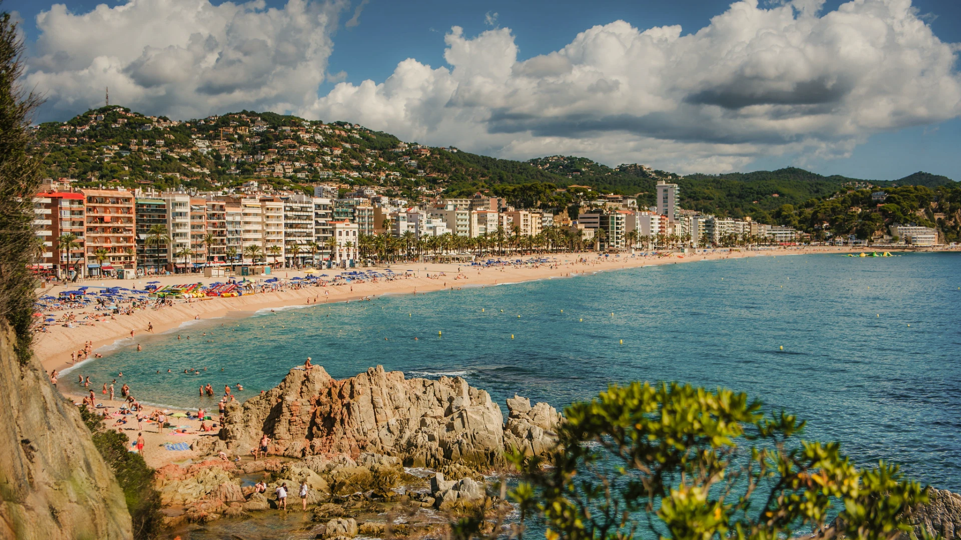 Playa con edificios y monta&ntilde;as al fondo, cielo nublado y personas disfrutando del mar.