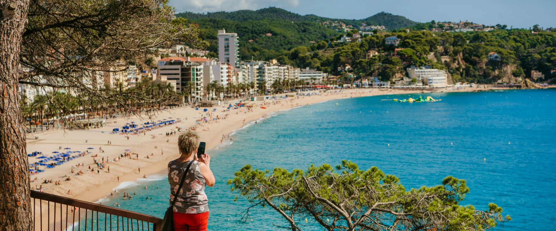 Persona toma una foto de una playa con edificios, vegetaci&oacute;n y colinas al fondo.