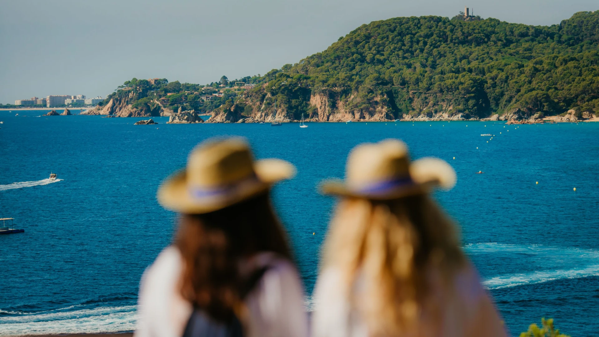 Dos personas con sombrero miran el mar y un paisaje de colinas con &aacute;rboles al fondo.