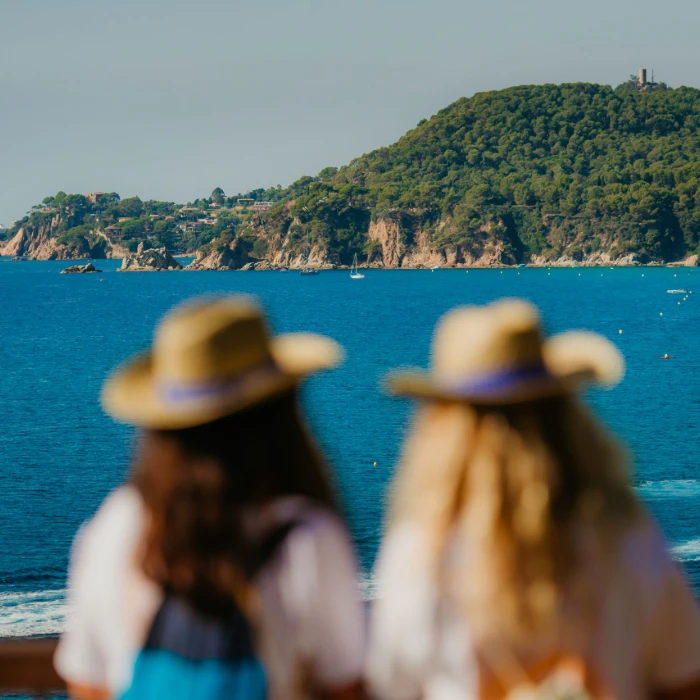 Dos personas con sombreros miran un paisaje mar&iacute;timo con una colina verde al fondo.