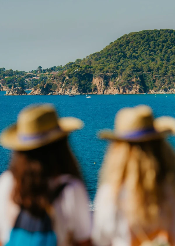 Dos personas de espaldas ven el mar y una isla boscosa, llevan sombreros y camisas blancas.