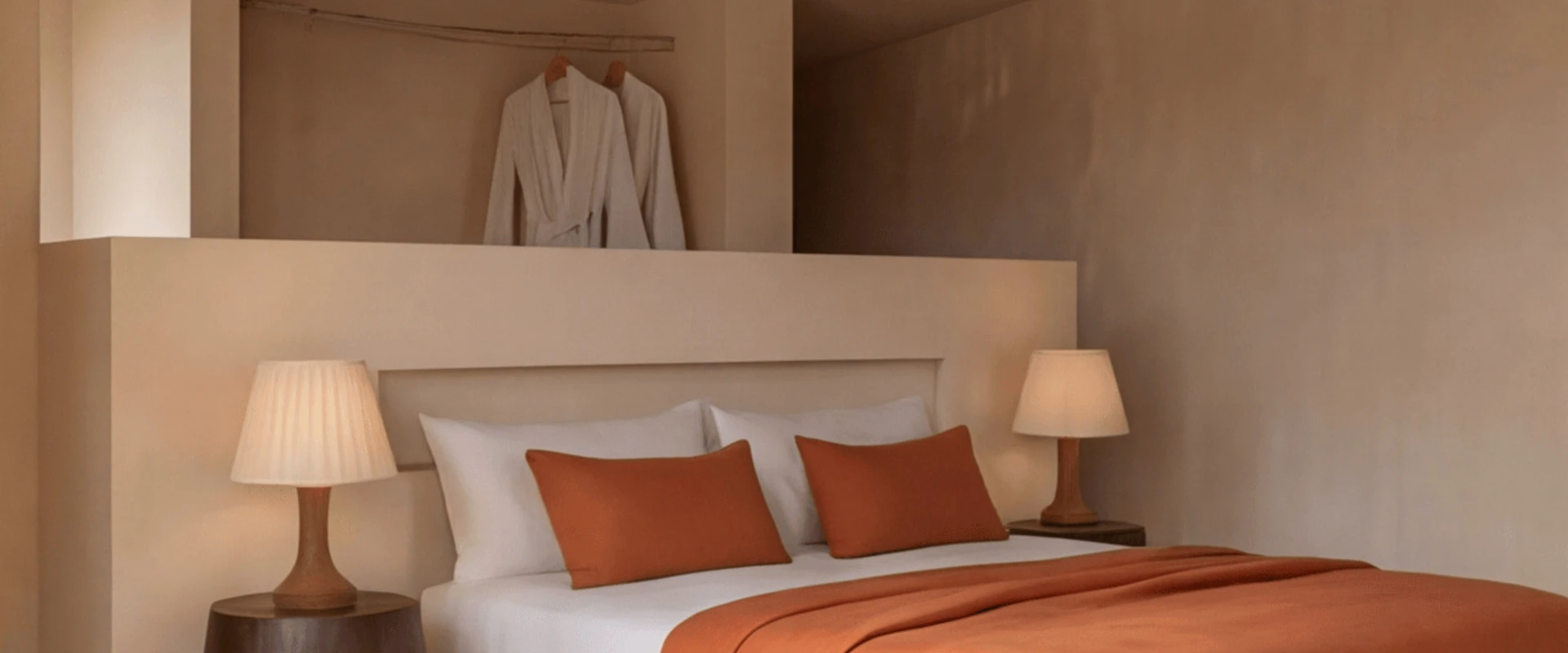 Habitaci&oacute;n con cama doble, almohadas y mantas naranjas, l&aacute;mparas y batas de ba&ntilde;o colgando.