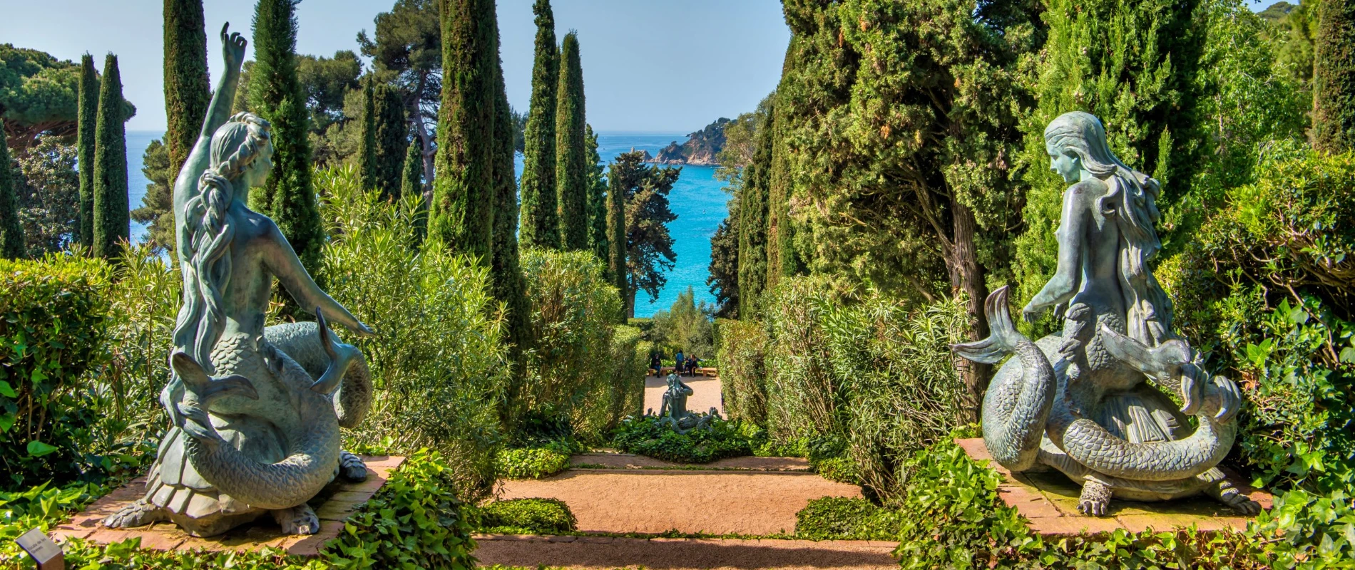Estatuas de sirenas en un jard&iacute;n rodeado de cipreses con vista a un mar azul.