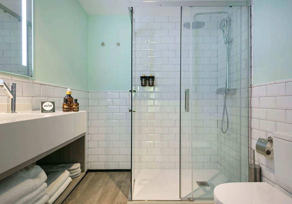 Ducha moderna con mampara de vidrio, azulejos blancos y lavabo con toallas