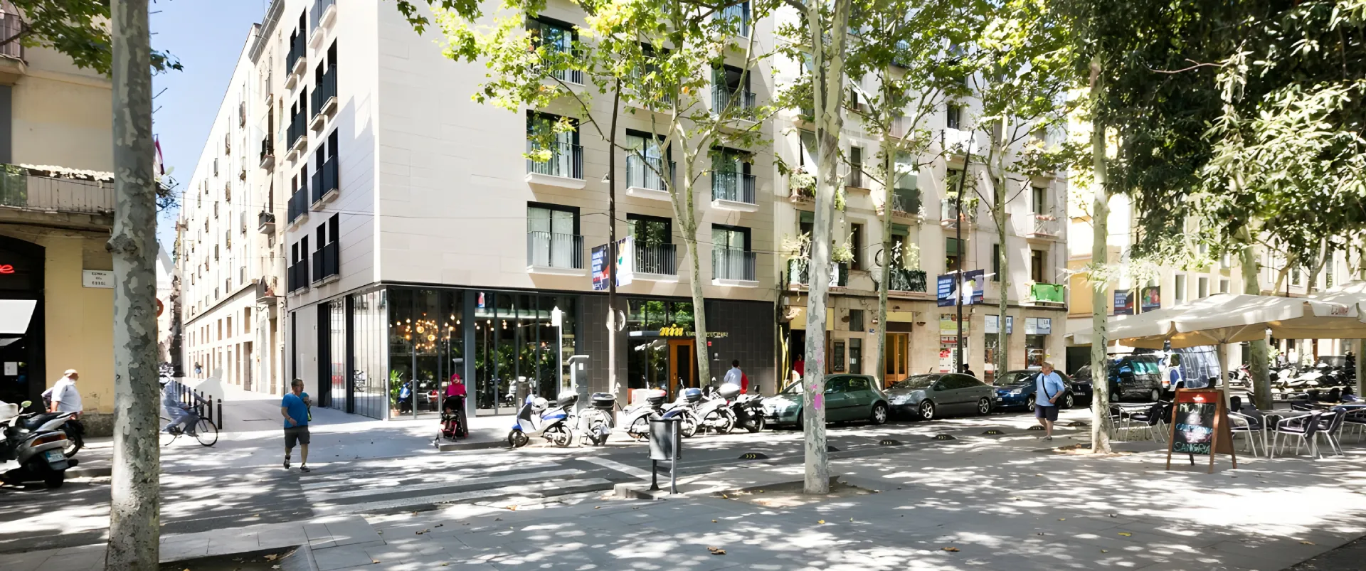 Calle urbana con edificios, &aacute;rboles y motocicletas aparcadas al sol